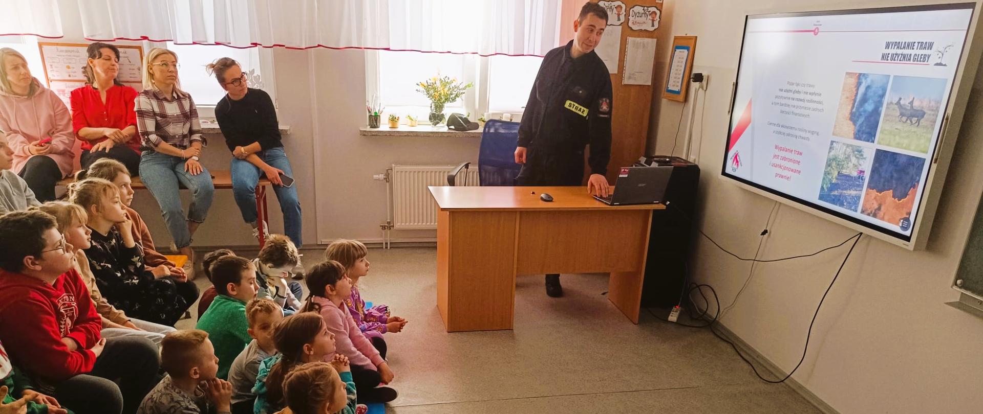 Zdjęcie przedstawia funkcjonariusza Komendy Powiatowej Państwowej Straży Pożarnej w Gołdapi, który przedstawia uczniom Szkoły Podstawowej w Pogorzeli informacje na temat kampanii społecznej Państwowej Straży Pożarnej „Stop Pożarom Traw”, m.in.: niebezpieczeństw jakie niesie za sobą wypalania traw oraz szkodliwości tego zjawiska dla społeczeństwa, zwierząt i środowiska, a także konsekwencji prawnych jakie spotykają tych, którzy mimo wszystko ryzykują i nielegalnie wypalają trawy