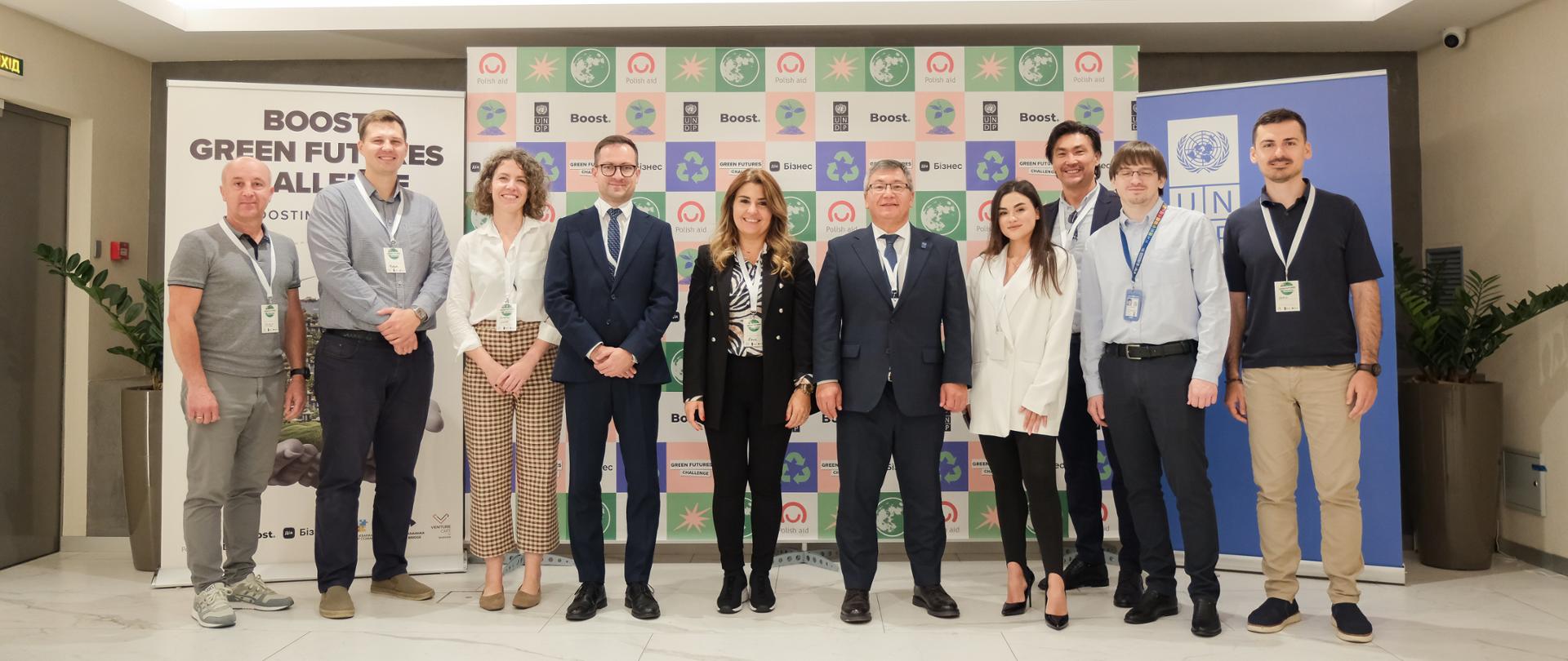 Grupa kobiet i mężczyzn pozuje na tle rollerów z logo Polska pomoc i UNDP Boost