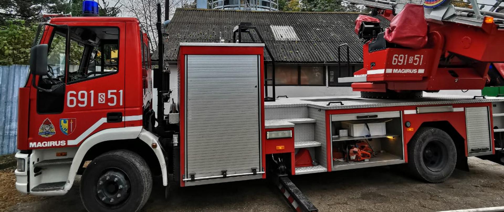 Drabina pożarnicza na samochodzie, rozłożona do górnej części silosu na trociny, który stoi za budynkiem. Przed drabiną leży rozwinięty wąż strażacki.