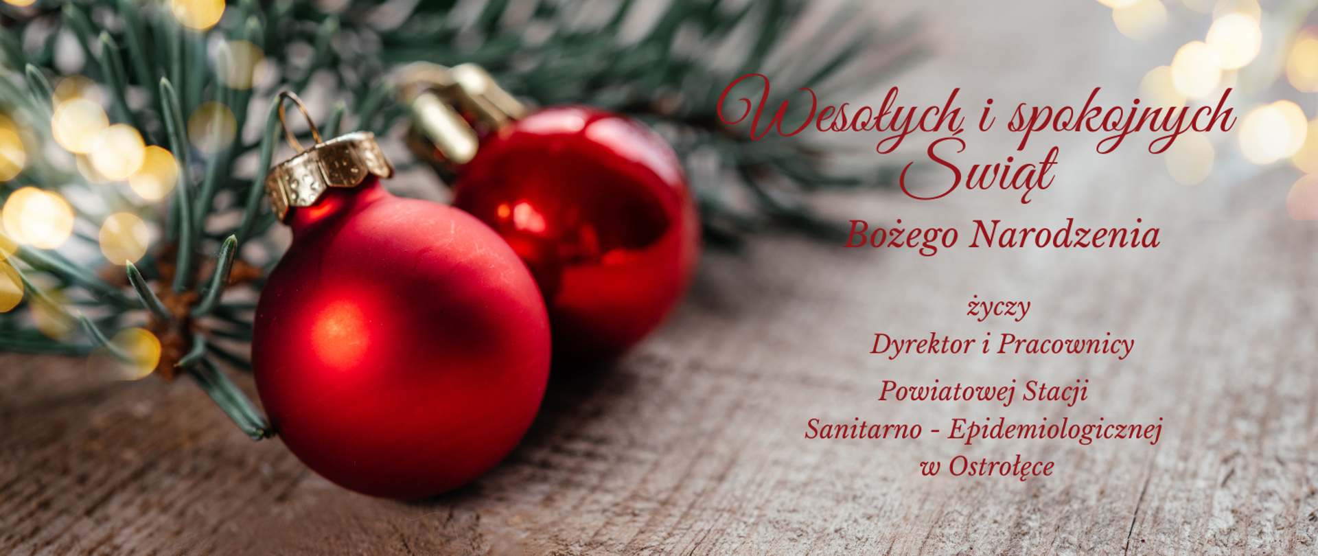 Na tle drewnianego stołu z lewej strony znajdują się dwie czerwone bombki ze złotymi zawieszkami. W lewym rogu za bombkami znajduje się zielona gałązka iglasta i plamy żółtych światełek. Z prawej strony znajduje się czerwony napis "Wesołych i spokojnych świąt Bożego Narodzenia życzy Dyrektor i Pracownicy Powiatowej Stacji Sanitarno-Epidemiologicznej w Ostrołęce". W prawym górnym rogu plamy żółtych światełek.