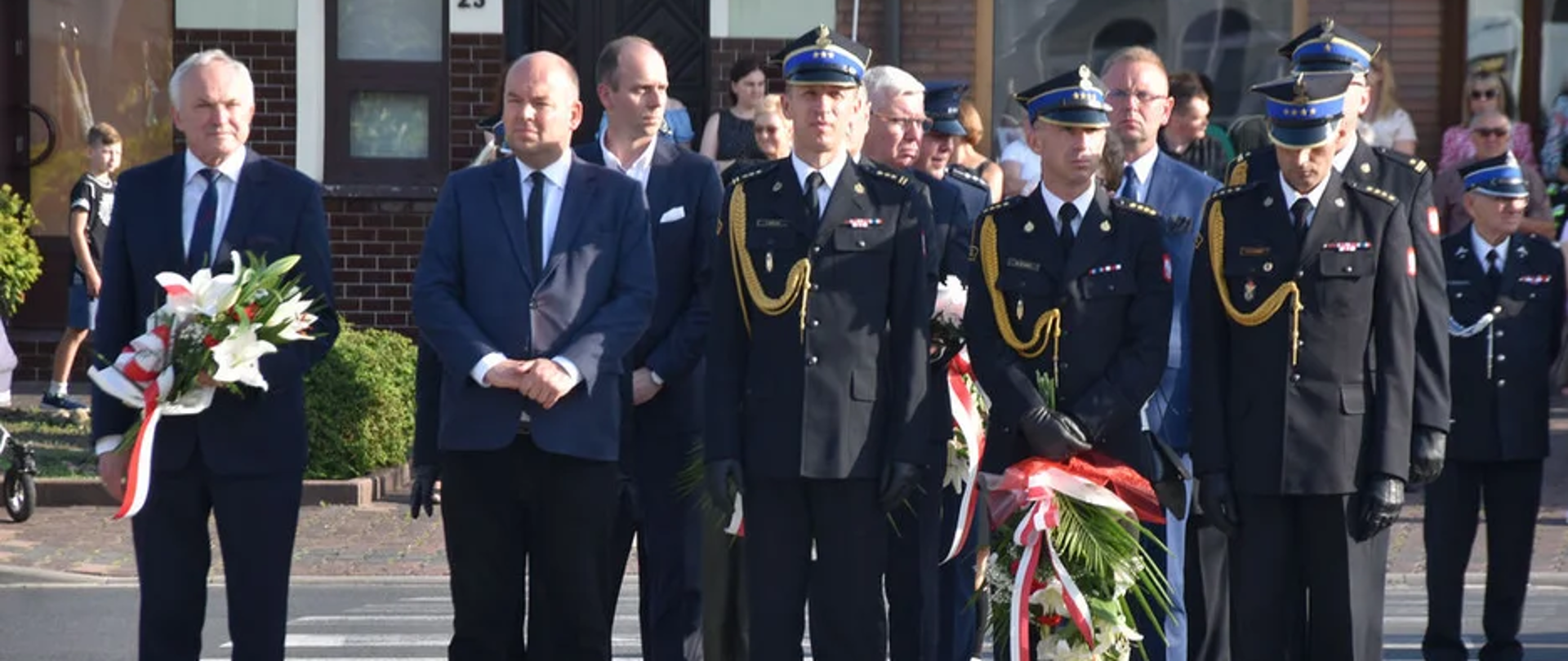 delegacja złożona ze strażaków i mężczyzn stoi z wieńcami , w tle grupa ludzi