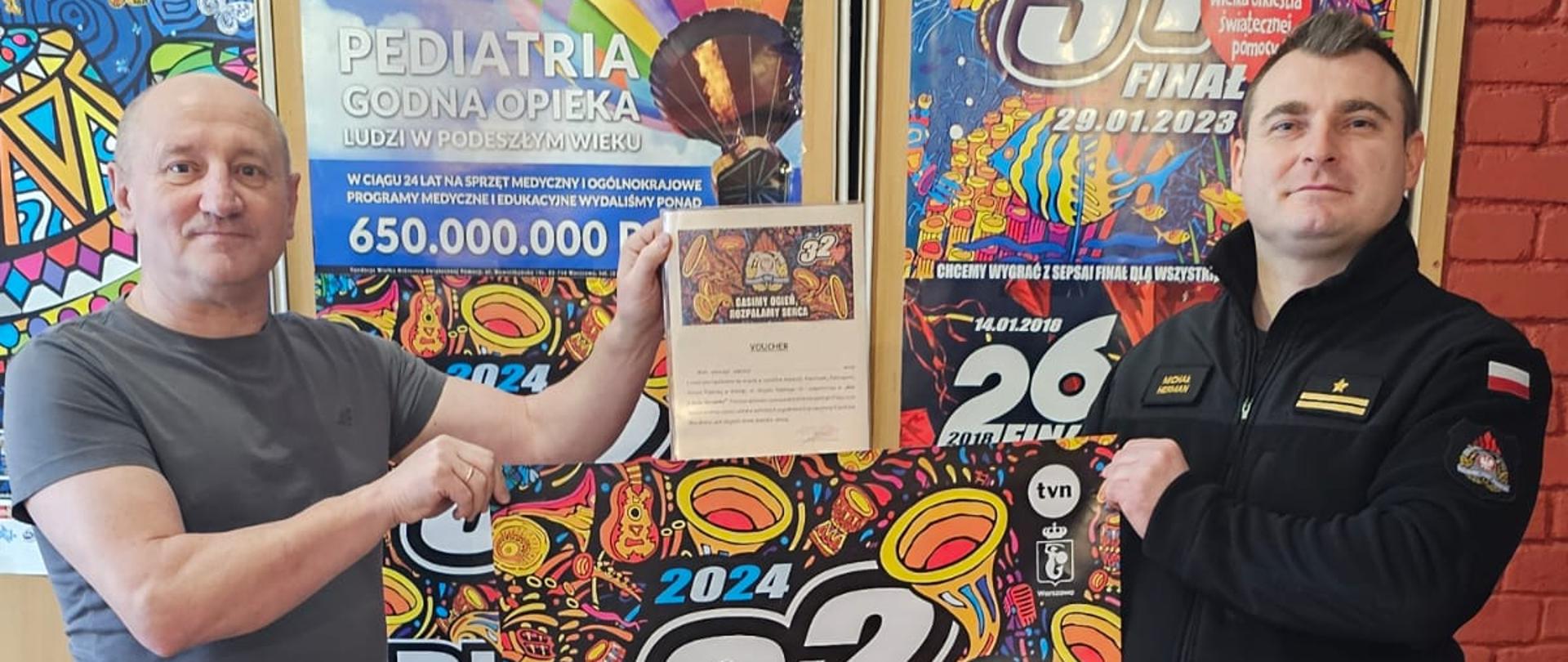 Zdjęcie przedstawia moment przekazania przez Zastępcę Komendanta Powiatowego Państwowej Straży Pożarnej w Gołdapi mł. bryg. Michała Herman przedstawicielowi sztabu Wielkiej Orkiestry Świątecznej Pomocy w Gołdapi voucher przeznaczony na licytację podczas 32. Finału WOŚP, który umożliwi darczyńcom wizytę w siedzibie gołdapskich strażaków i uczestnictwo „w dniu z życia Strażaka”