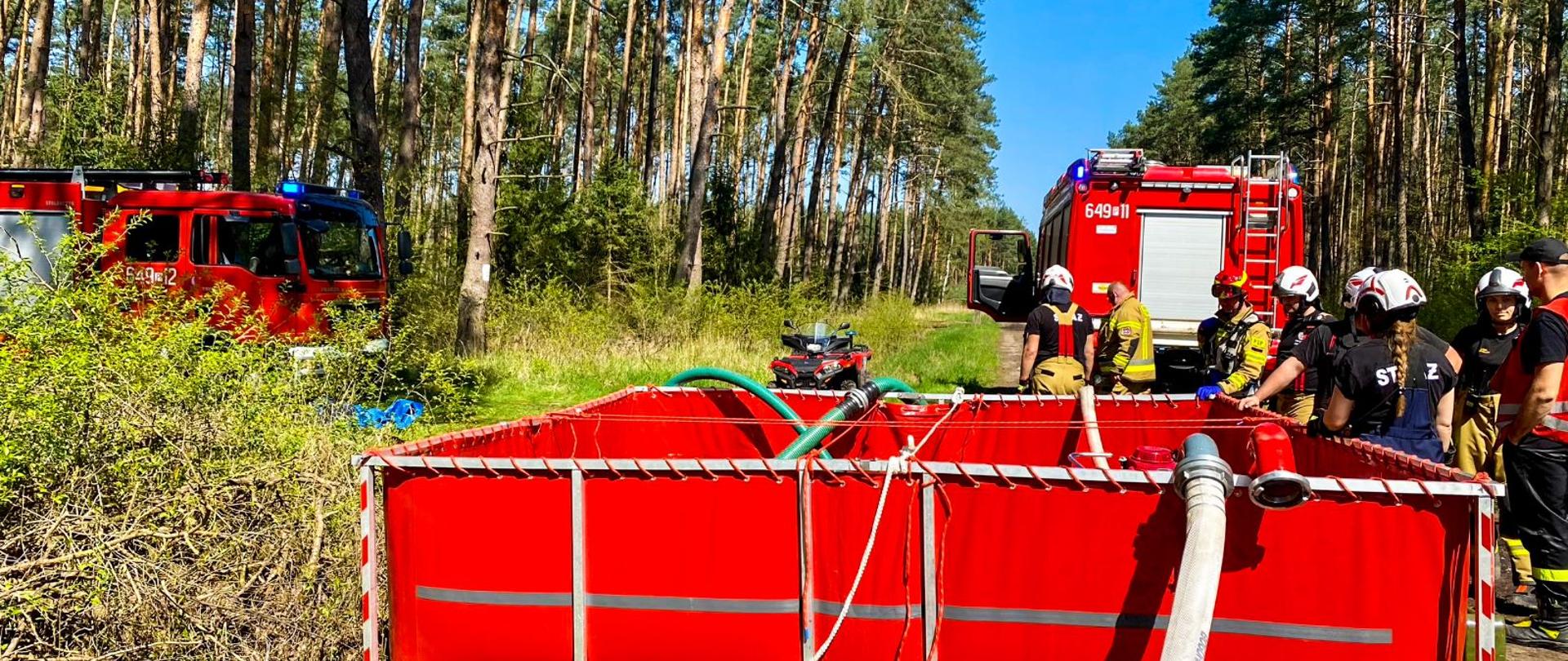 Zdjęcie przedstawia punkt czerpania wody, stojących strażaków przy zbiorniku. W tle widzimy dwa pojazdy gaśnicze oraz quad. 