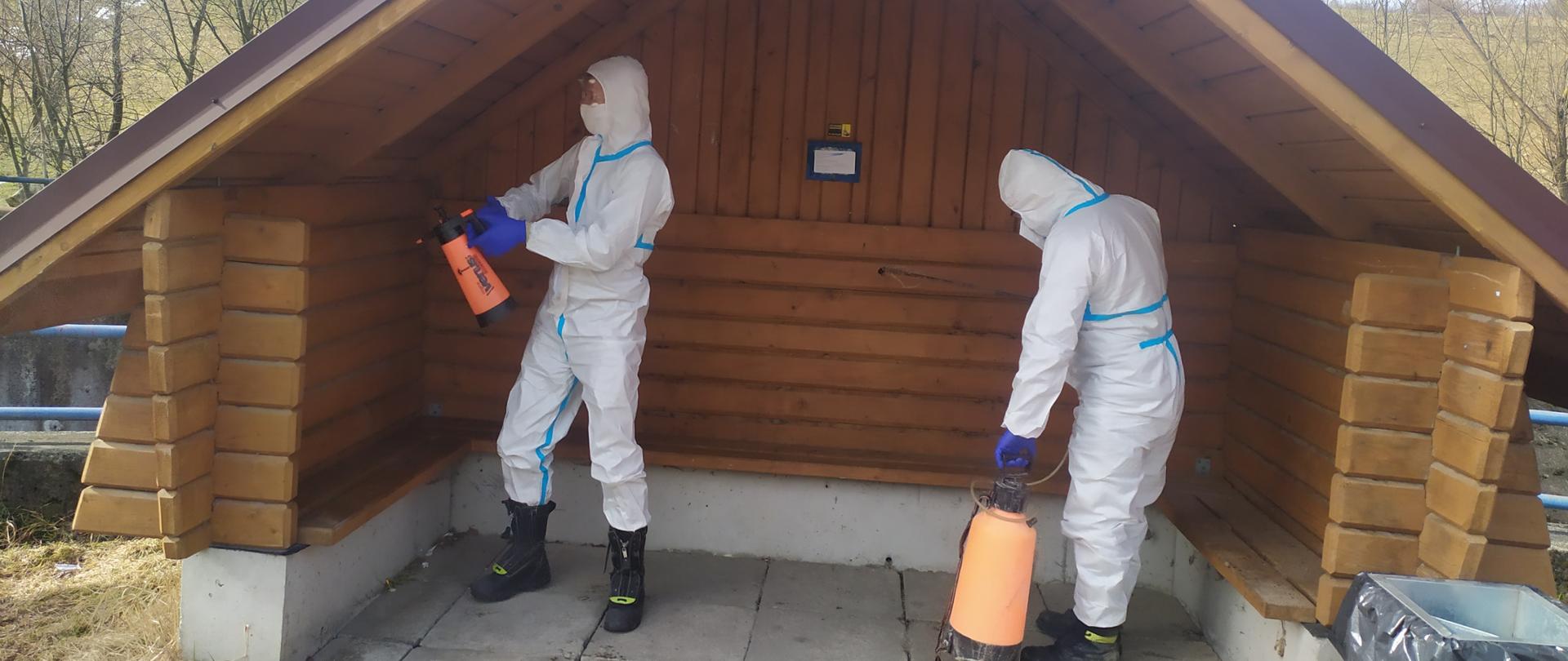 Strażacy w specjalnych kombinezonach, opryskiwaczami ręcznymi dezynfekują wiatę przystanku. 