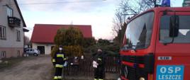 Na zdjęciu po lewej stronie na pierwszym planie przód samochodu strażackiego z napisem na drzwiach OSP Leszcze. Na środku zdjęcia dwóch strażaków w umundurowaniu bojowym i koszarowym, jeden z nich trzyma jasną kartkę. Strażacy stoją na tle drewnianego płotu. Na drugim planie drzewa i krzewy ozdobne oraz fragmenty dwóch budynków oraz wjazd na posesję.