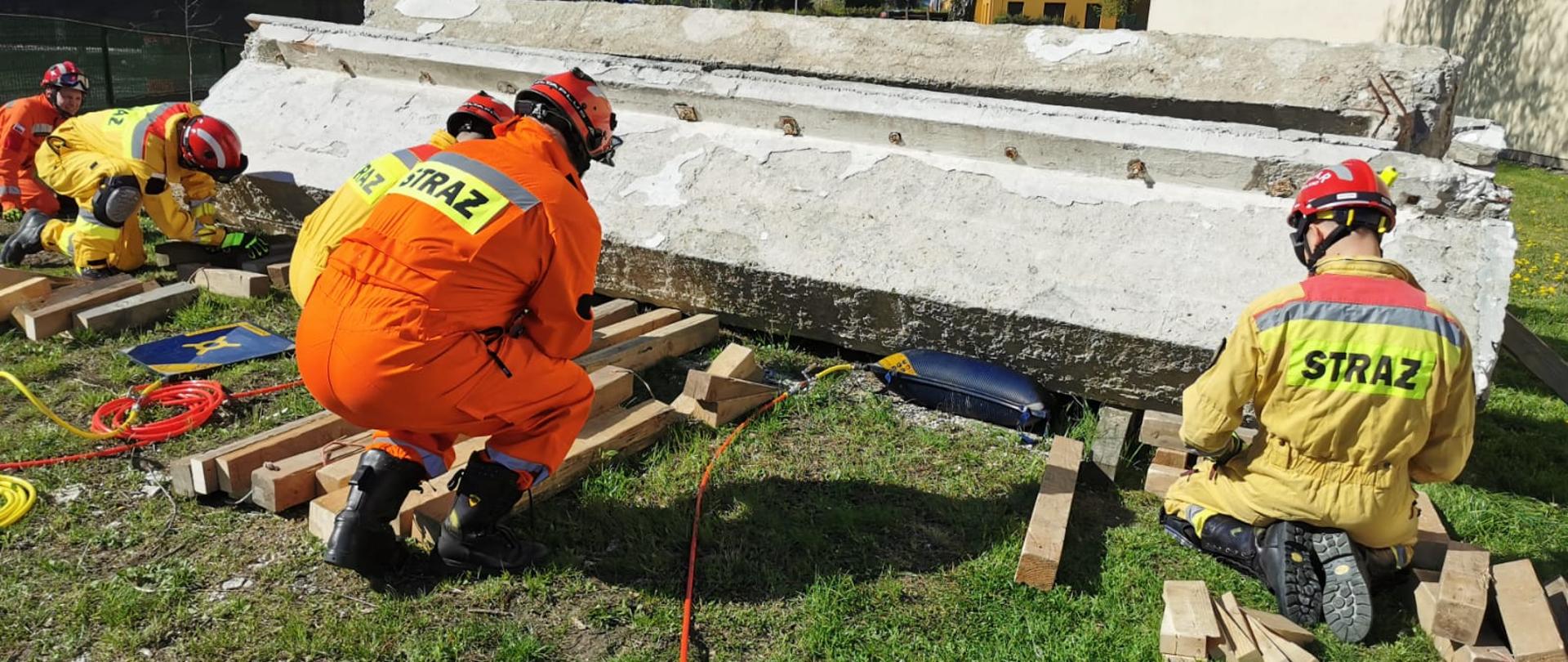 Na zdjęciu jest pięciu ratowników SGPR ubranych w kombinezony i kaski ochronne. Ratownicy unoszą dużą konstrukcję żelbetonową za pomocą czarnych poduszek pneumatycznych.