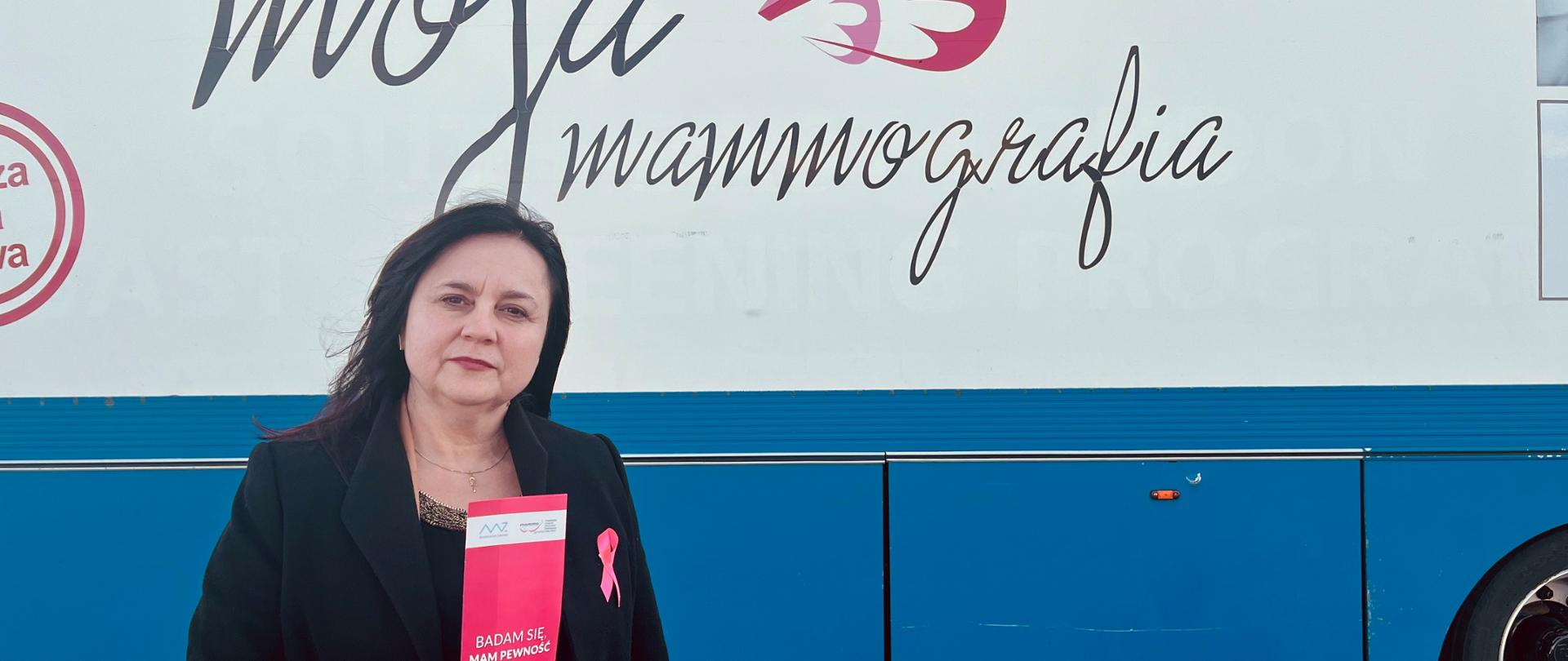 Państwowy Powiatowy Inspektor Sanitarny w Sochaczewie Beata Fergińska promuje badania profilaktyczne piersi. Trzyma w ręku ulotkę, a do płaszcza przypiętą ma różową wstążeczkę. W tle mammobus.