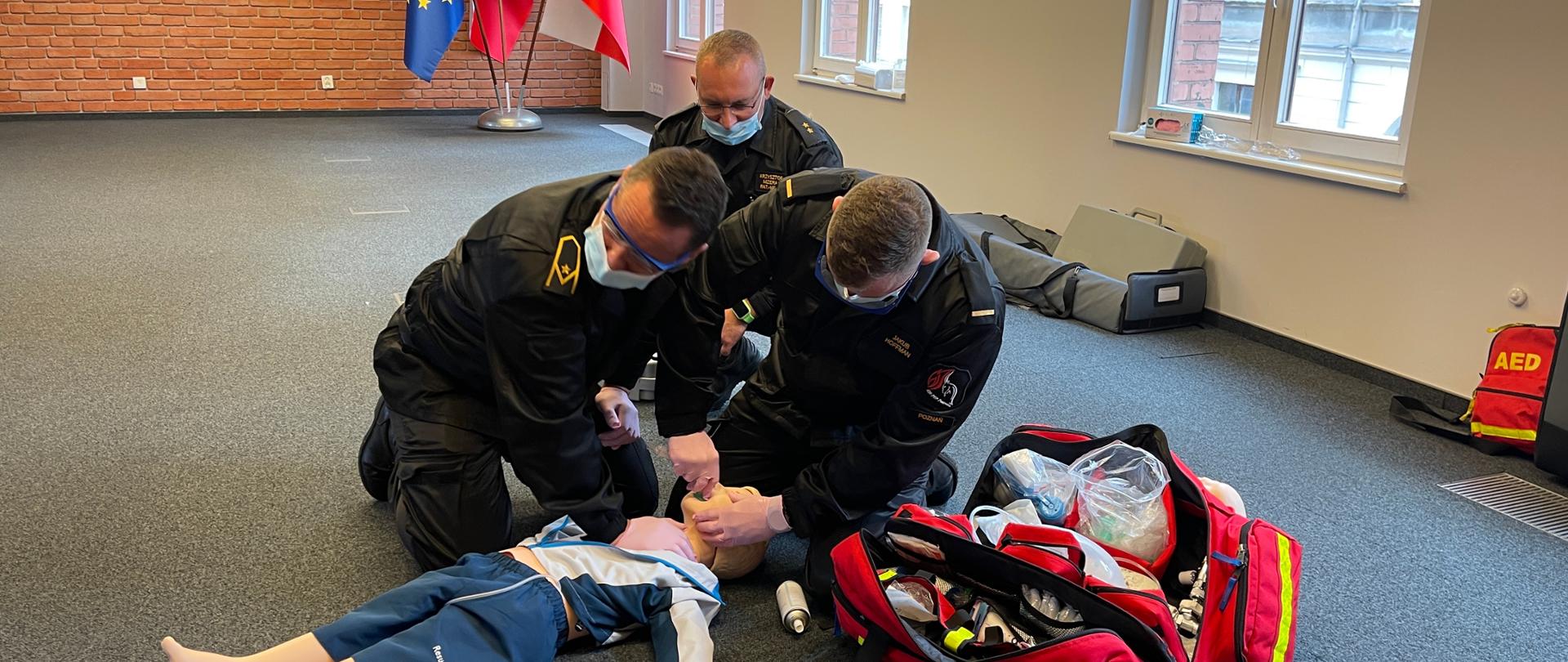 Jeden strażak udrażnia drogi oddechowe fantomowi dziecka, podczas gdy drugi strażak jedną ręką uciska klatkę piersiową tego fantoma.