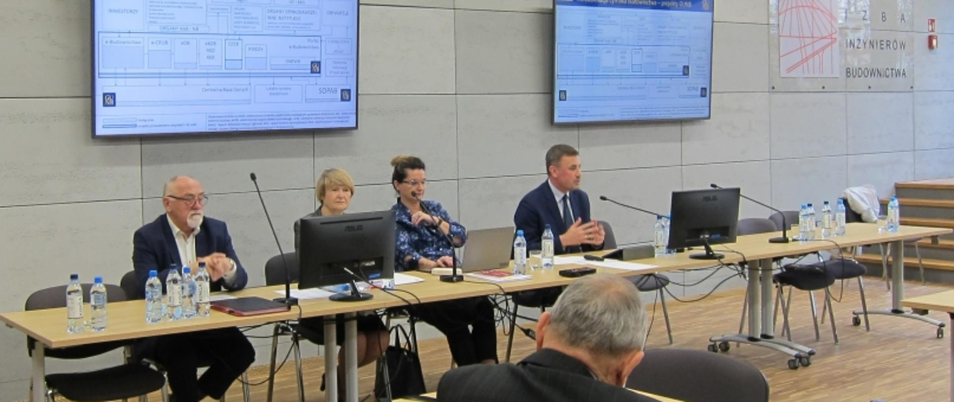 Cztery osoby siedzą przy stole konferencyjnym z mikrofonami; za nimi dwa ekrany i napis „Inżynierów Budownictwa”.