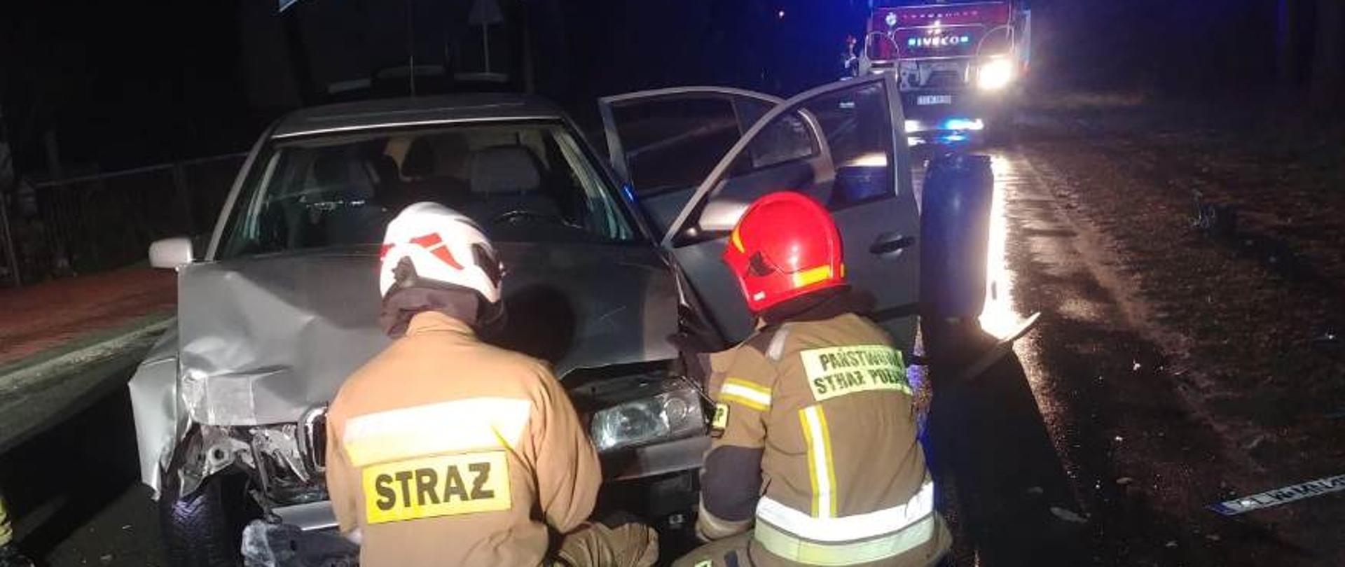 Droga wojewódzka numer 742 w miejscowości Kluczewsko, samochód osobowy Skoda Octavia blokuje ruch w obu kierunkach, samochód ma uszkodzony przód, dwóch strażaków ratowników podnosi maskę samochodu celem odłączenia akumulatora w pojeździe, na drugim planie samochód pożarniczy zabezpieczający miejsce zdarzenia. 