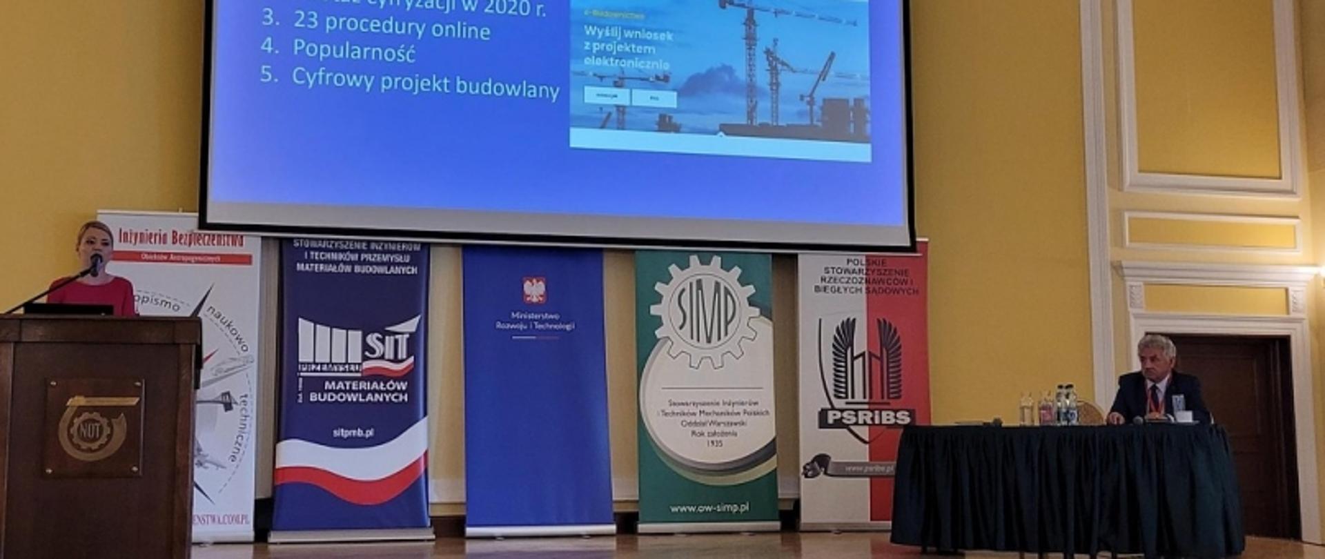 Sala konferencyjna z prezentacją dotyczącą cyfryzacji budownictwa, prelegentem przy mównicy i ekranem z agendą zawierającą punkty o procedurach online i projekcie cyfrowym.