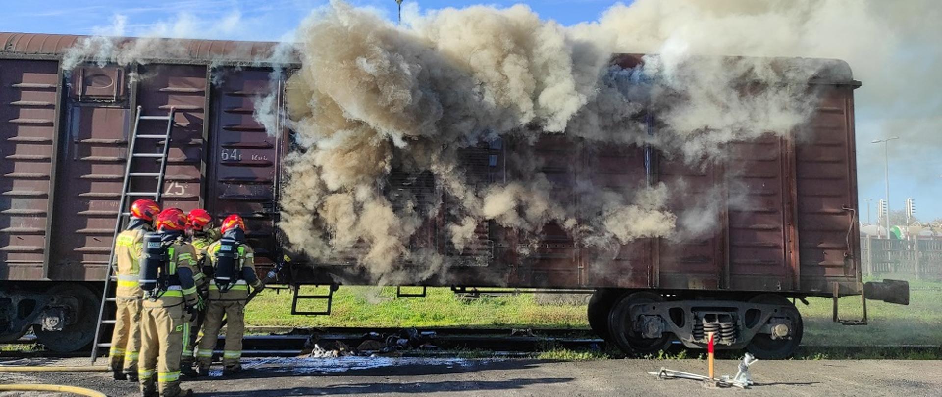 Na zdjęciu wagon z którego wydobywa się brunatny dym. Obok stoją strażacy w mundurach i czerwonych hełmach.