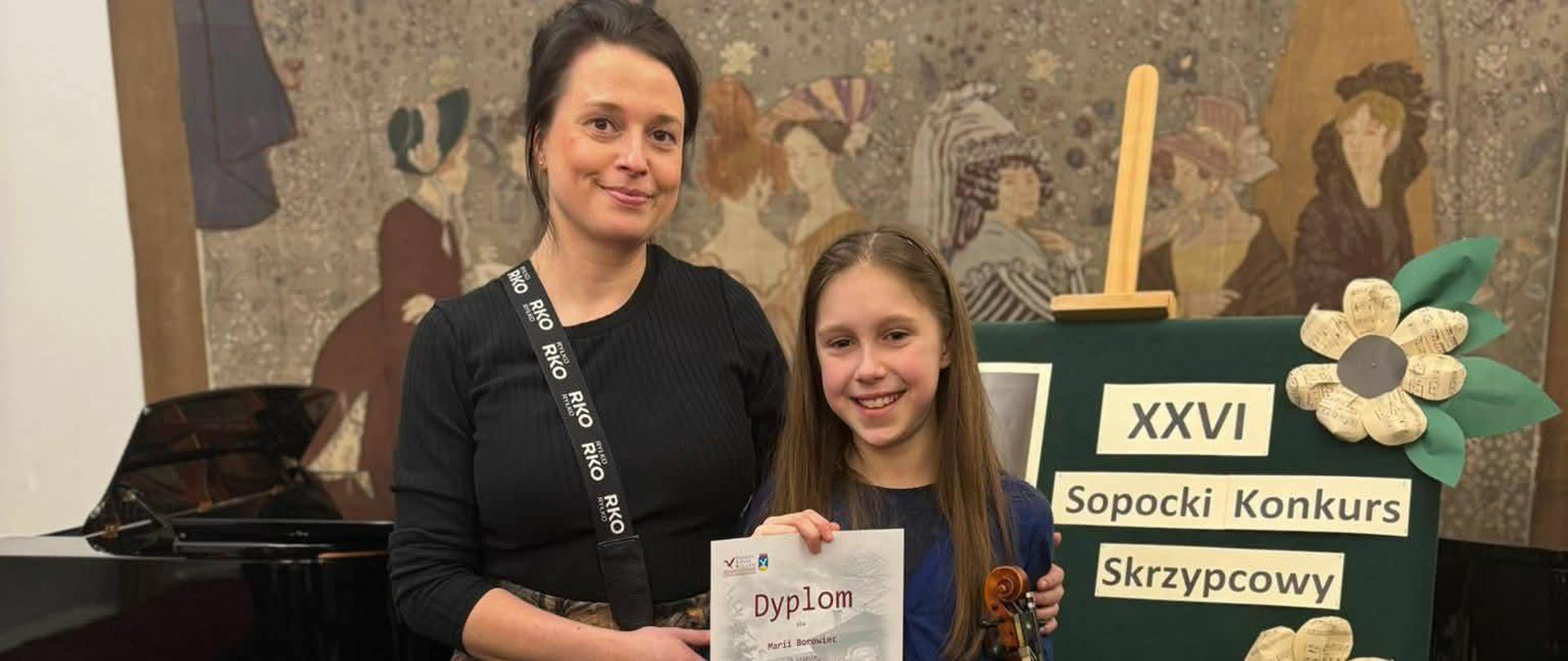 Na zdjęciu nauczycielka Agata Parzych wraz z uczennicą. Pozują na stojąco. W tle sztaluga z napisem "XXVI Sopocki Konkurs Skrzypcowy"