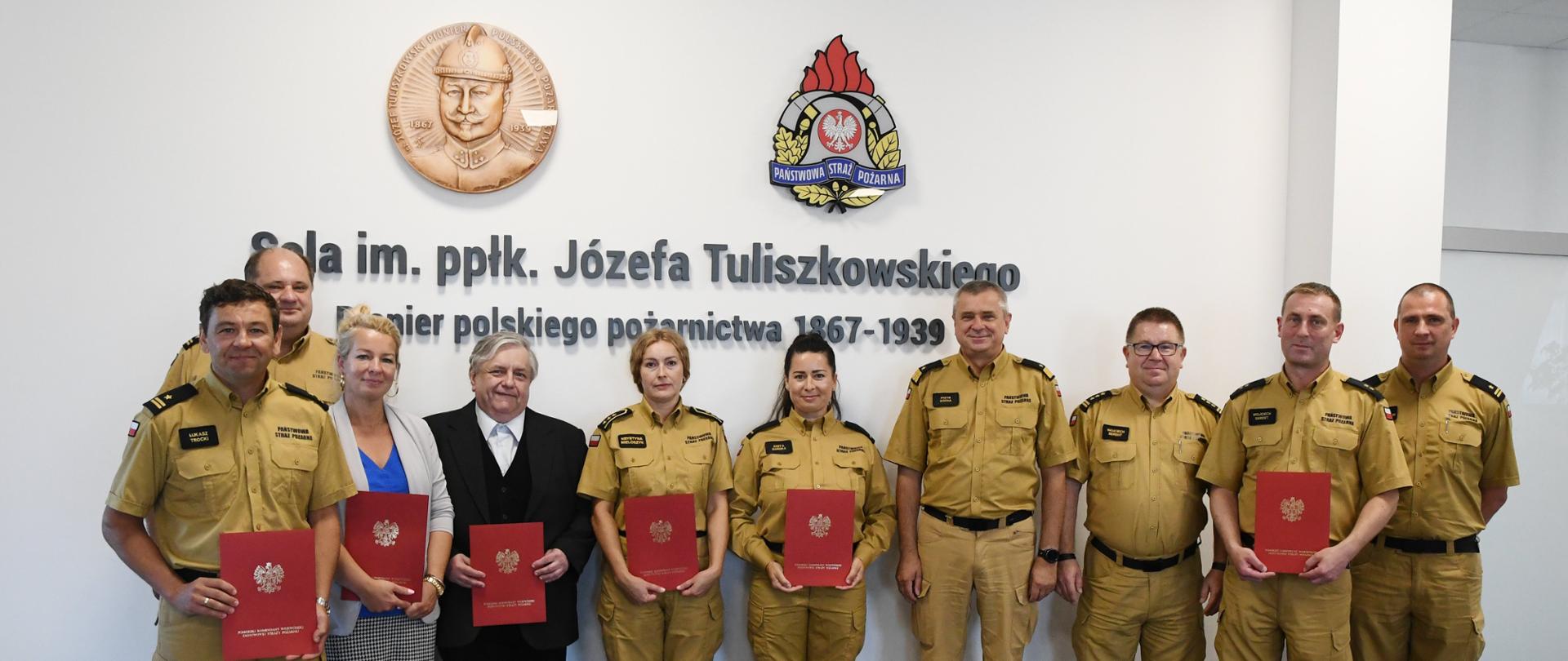 Strażacy w mundurach musztardowych oraz pracownicy cywilni trzymający czerwone teczki stoją wspólnie z kierownictwem Komendy Wojewódzkiej Państwowej Straży Pożarnej w Gdańsku za nimi na ścianie jest napis sala imieniem podpułkownika Józefa Tuliszkowskiego pioniera polskiego pożarnictwa nad napisem wisi logo Państwowej Straży Pożarnej oraz popiersie funkcjonariusza. 