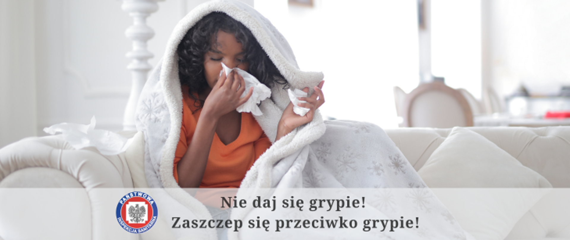 Zdjęcie przestawia kobietę siedząca na jasnej sofie przykrytej jasnym kocem, w ręku trzyma chusteczkę którą wyciera nos