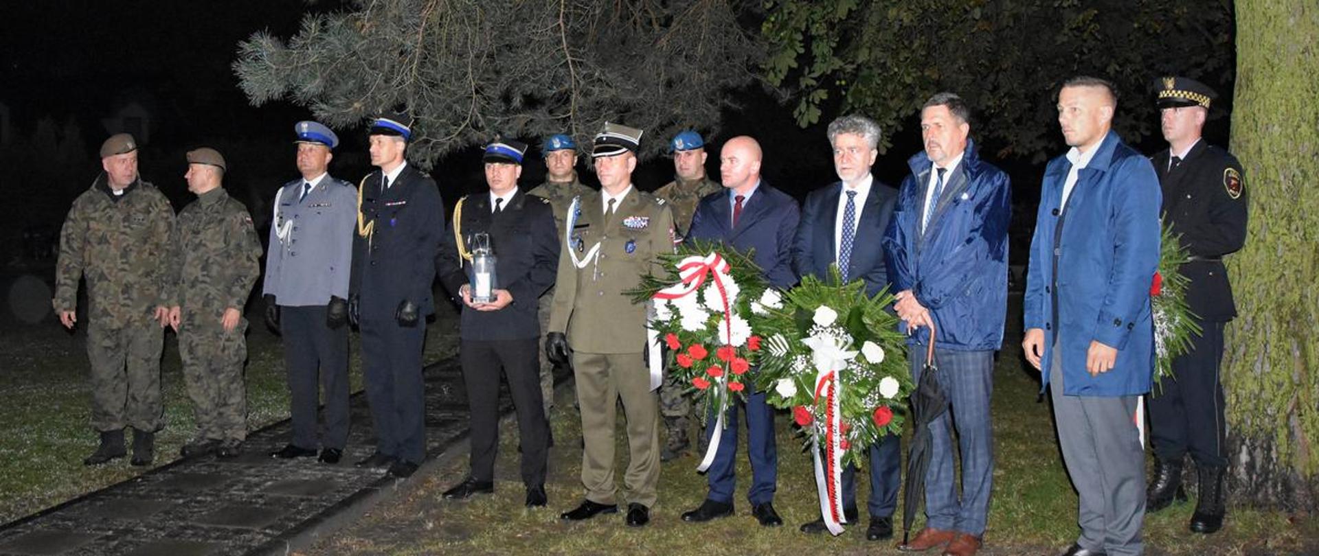 Zdjęcie przedstawia stojących w jednym rzędzie przedstawicieli służb mundurowych (Policja, Straż Pożarna i Wojsko) oraz przedstawicieli władz terenowych i samorządowych., którzy składając wieńce upamiętnili 82 rocznicę wybuchu II Wojny Światowej.