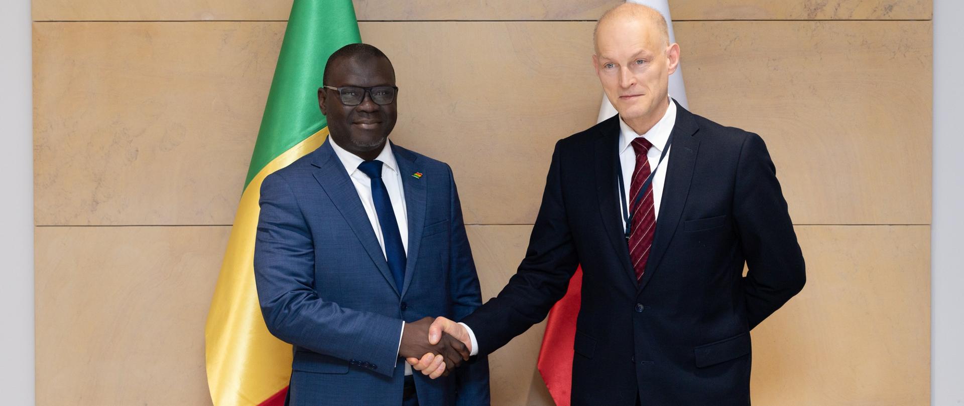 Two men in suits shake hands during a diplomatic meeting, standing in front of the flags of Senegal and Poland against a wall featuring the emblem of the Republic of Poland.