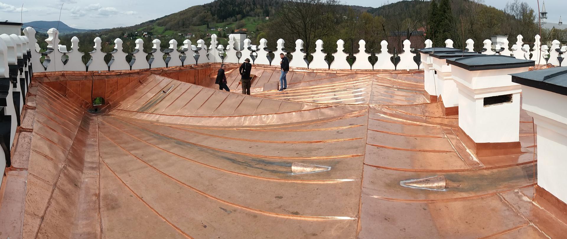 Panoramiczny widok nowego miedzianego poszycia dachowego otoczonego attyką. Po prawej widoczne 3 duże kominy.
Prace podczas projektu poza wymianą poszycia dotyczyły także remontu rozbudowanych detali ściany attykowej.