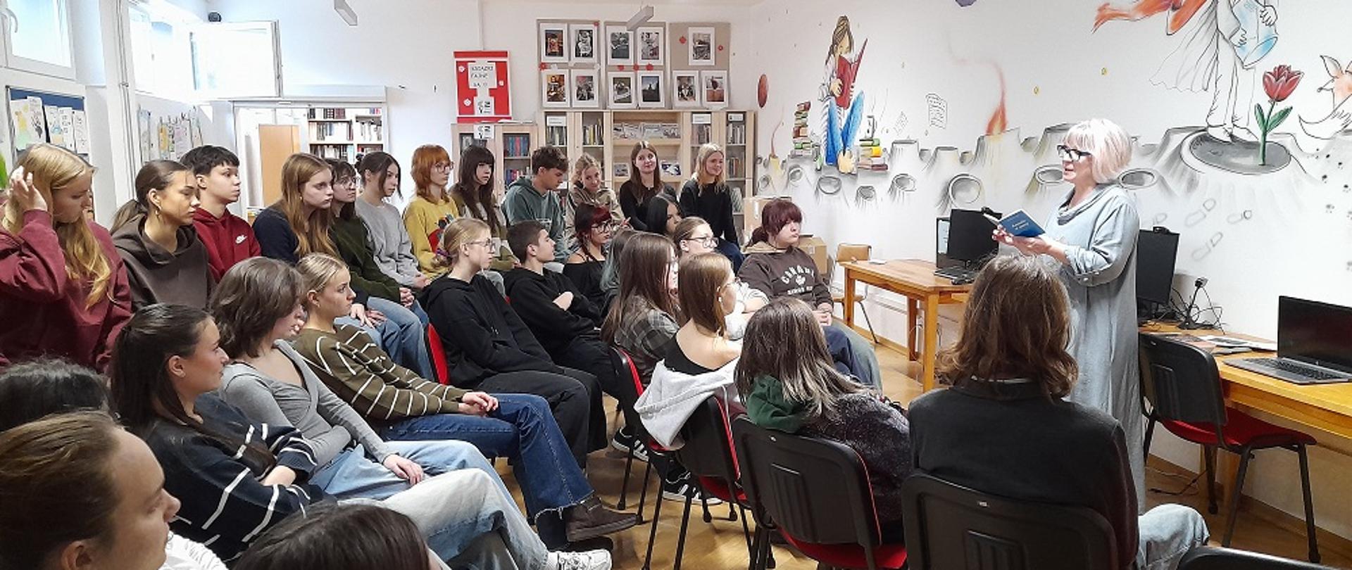 Uczniowie siedzą w bibliotece, słuchają nauczyciela w czytaniu autorskim