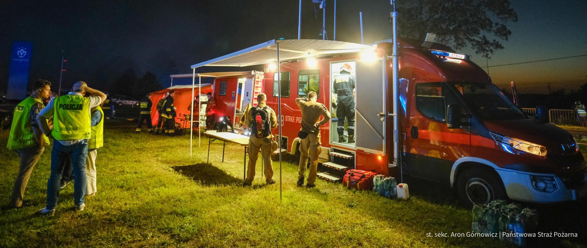 Nocna akcja służb ratunkowych – mobilne centrum dowodzenia straży pożarnej z wysuniętym zadaszeniem, otoczone przez strażaków i policjantów w kamizelkach odblaskowych, prowadzących działania w terenie.