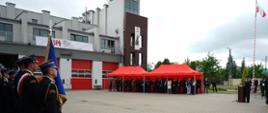 Na tle budynku po lewej stronie Sztandar Komendy Powiatowej Państwowej Straży Pożarnej w Lublińcu, w środkowej części pod baldachimem grupa zaproszonych gości. Po prawej stronie flaga państwowa wciągnięta na maszt.