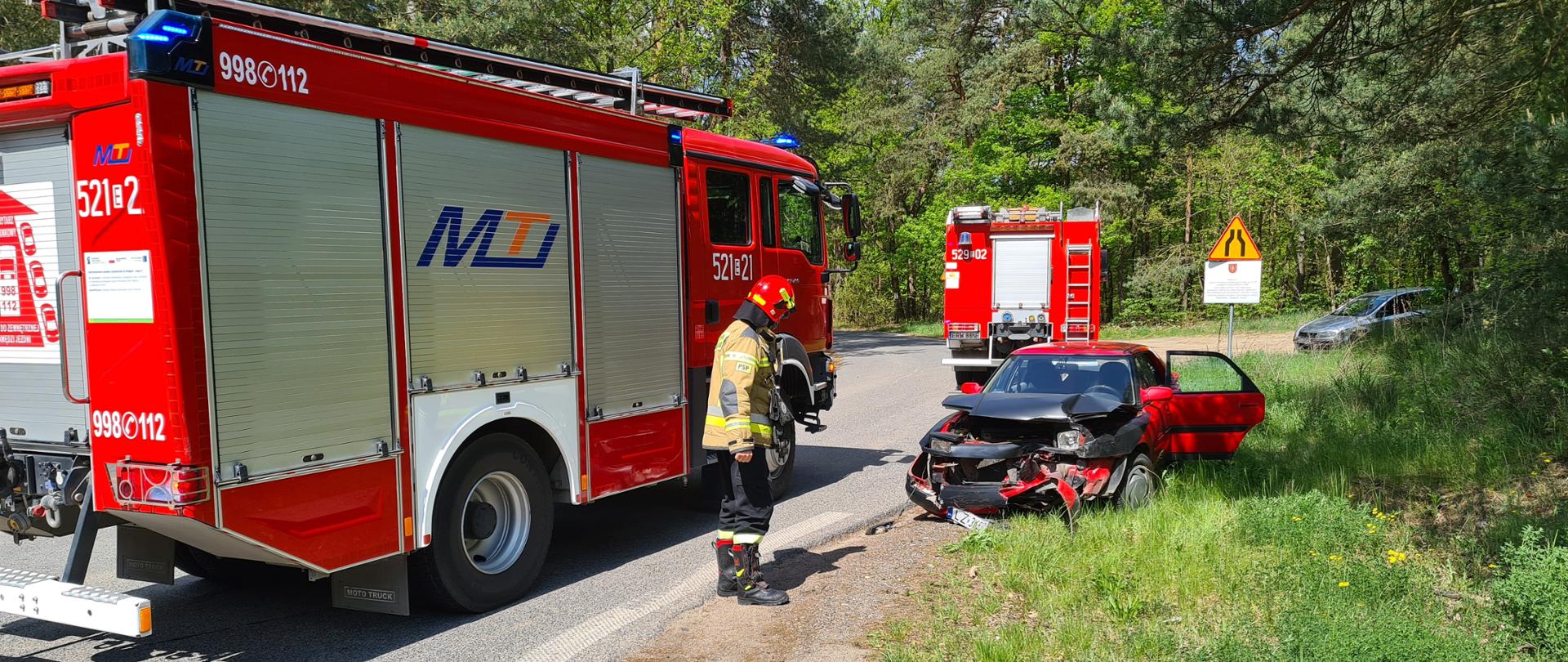 Zdjęcie przedstawia 2 samochody strażackie oraz 2 samochody osobowe oraz strażaka na drodze, a dookoła widać zielone drzewa/las. Na pierwszym planie znajduje się samochód bojowy strażacki, skierowany prawym bokiem do fotografa, przy samochodzie stoi strażak ubrany w strój strażacki oraz hełm i parzący na stojący przed nim czerwony samochód osobowy, ze skasowaną przednią maską w wyniku wypadku. Za samochodem widać tył drugiego samochodu strażackiego, znak drogowy oraz srebrny samochód osobowy.