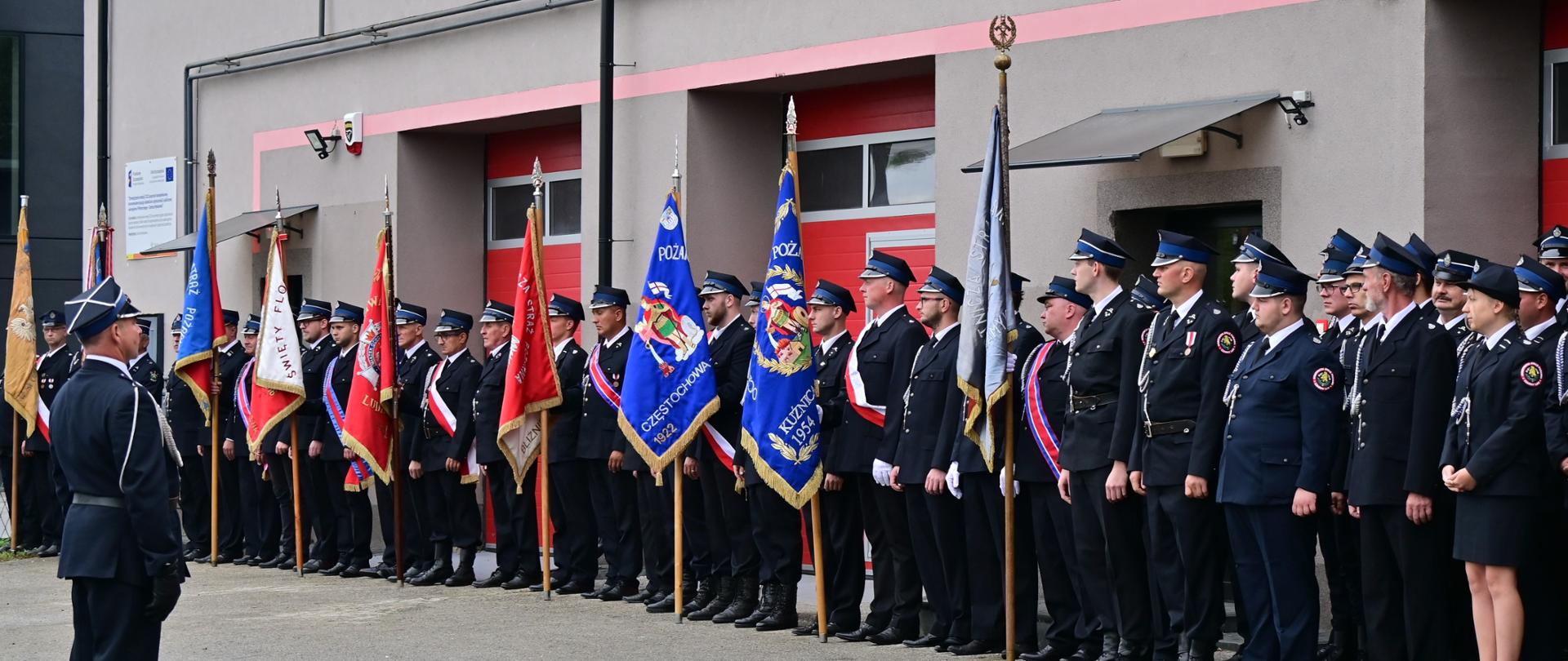 Poczty sztandarowe ustawione w rzędzie na tle budynku remizy OSP. Na budynku baner z napisem 100-lecie OSP Czarny Las 1923-2023