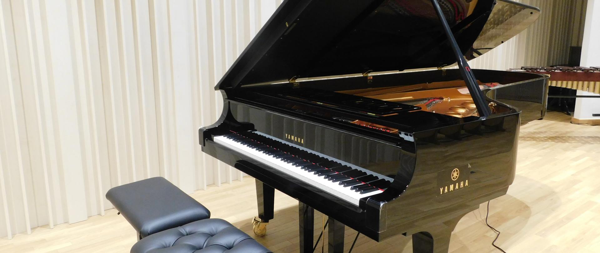 Na zdjęciu czarny fortepian z dwiema czarnymi ławami. Fortepian marki Yamaha. Fortepian ustawiony na drewnianej podłodze.
