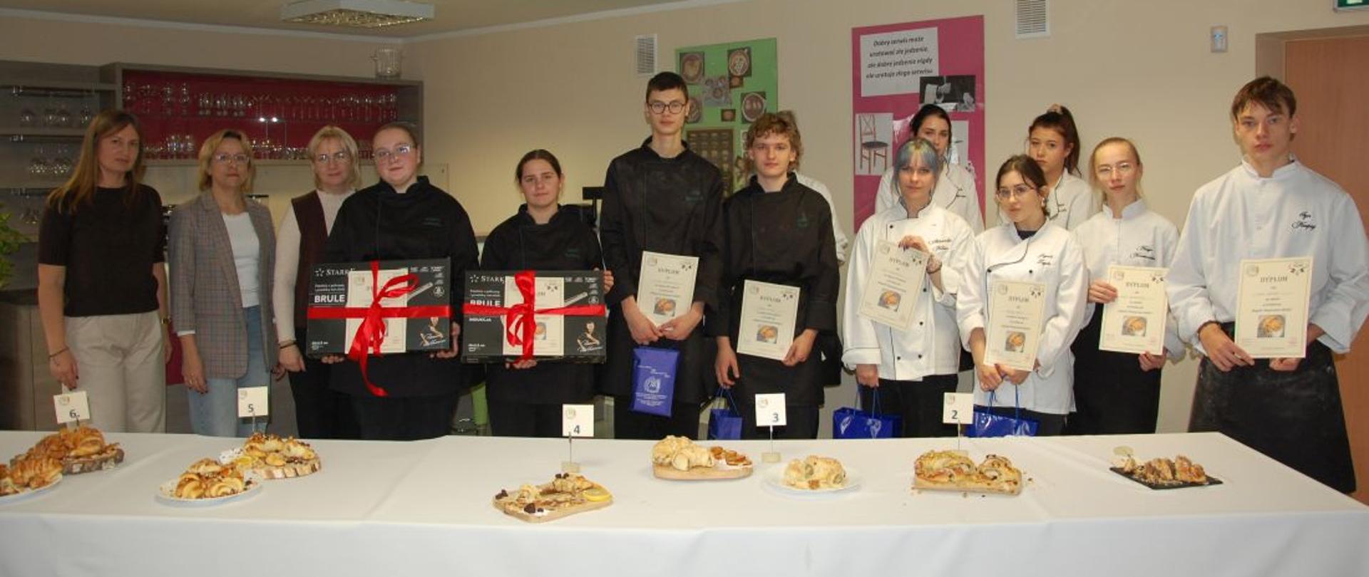 Uczniowie biorący udział w konkursie z organizatorami stoją za stołem zastawionym rogalami świętomarcińskimi, trzymają dyplomy i nagrody