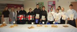 Uczniowie biorący udział w konkursie z organizatorami stoją za stołem zastawionym rogalami świętomarcińskimi, trzymają dyplomy i nagrody