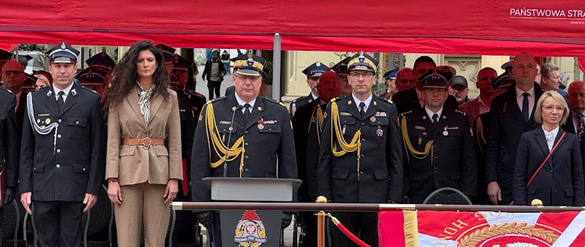 Zdjęcie przedstawia oficjalną, państwową uroczystość w Polsce, odbywającą się na świeżym powietrzu. Grupa osób, w tym umundurowani funkcjonariusze straży pożarnej oraz cywile, stoi w rzędzie pod zadaszeniem dużego, czerwonego namiotu eventowego.