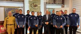 Zdjęcie grupowe kartuskich strażaków w nowych strojach sportowych po otrzymaniu listów gratulacyjnych od starosty. W tle sztandary KP PSP w Kartuzach