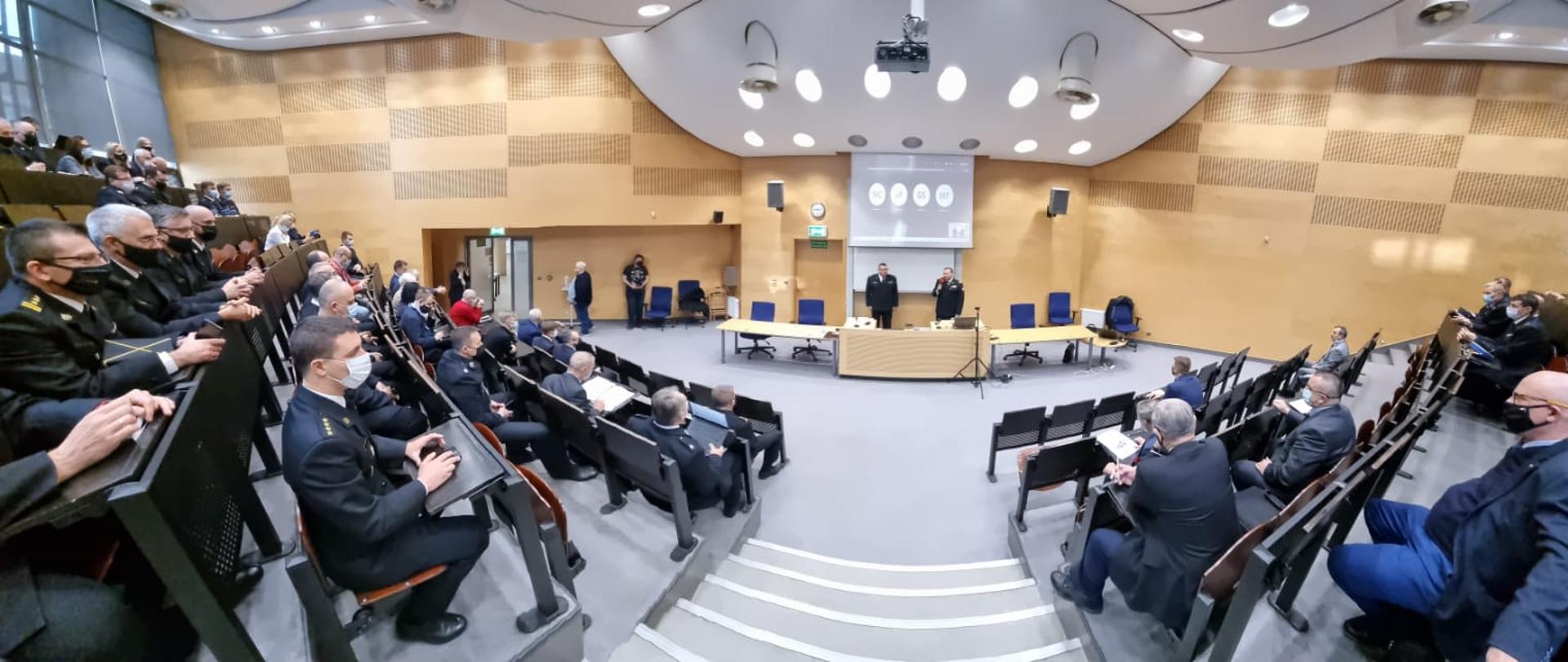 Widok na salę w której odbywa się konferencja. Widoczni siedzący na uli w maseczkach ochronnych uczestnicy, oraz w oddali dwóch prowadzących