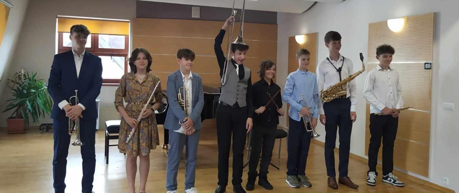 Ośmiu absolwentów sekcji instrumentów dętych stoją na scenie. W rękach trzymają instrumenty muzyczne. Za nimi fortepian oraz duży kwiatek.