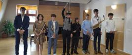 Ośmiu absolwentów sekcji instrumentów dętych stoją na scenie. W rękach trzymają instrumenty muzyczne. Za nimi fortepian oraz duży kwiatek.