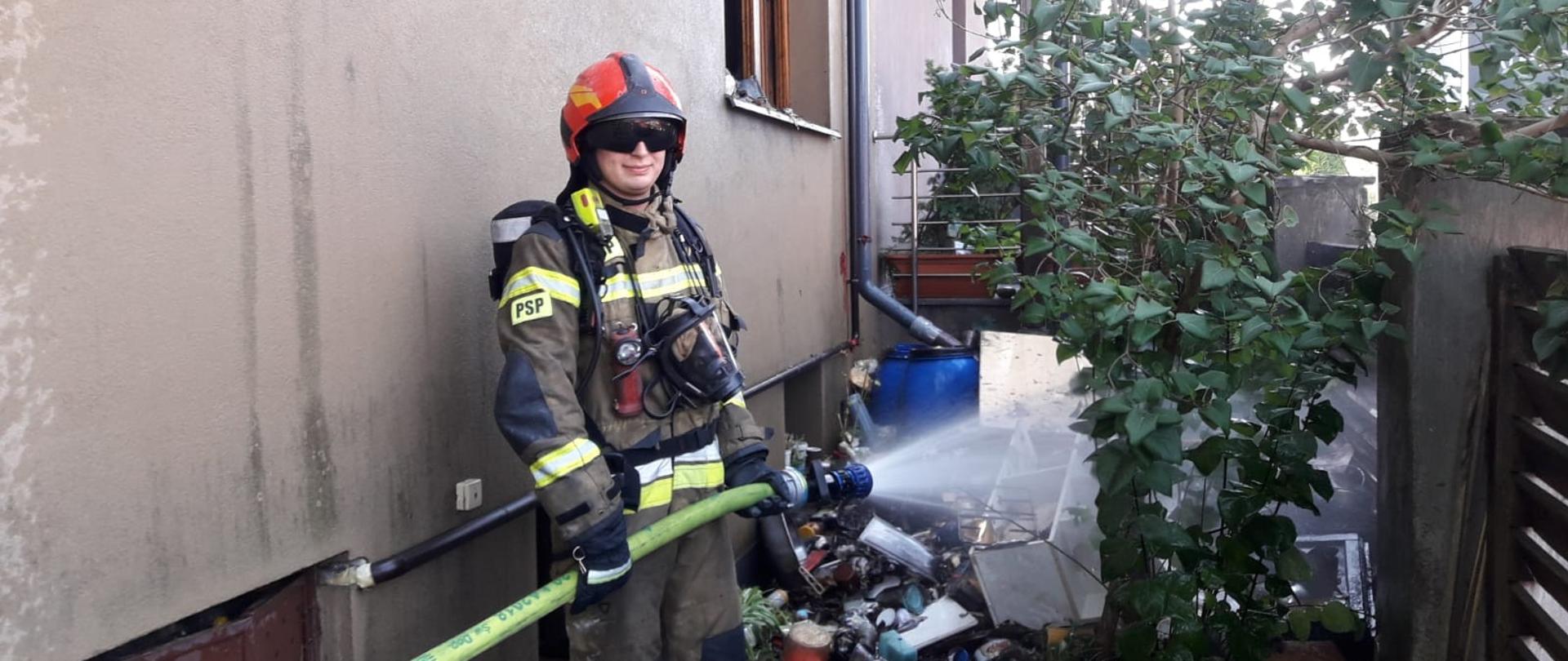 Strażak podaje prąd wody na spalone elementy przed budynkiem.