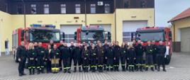 Druhny i druhowie OSP wraz z funkcjonariuszami PSP stoją przed garażami, za nimi stoją 3 pojazdy gaśnicze PSP. Za nimi widoczny jest budynek KP 