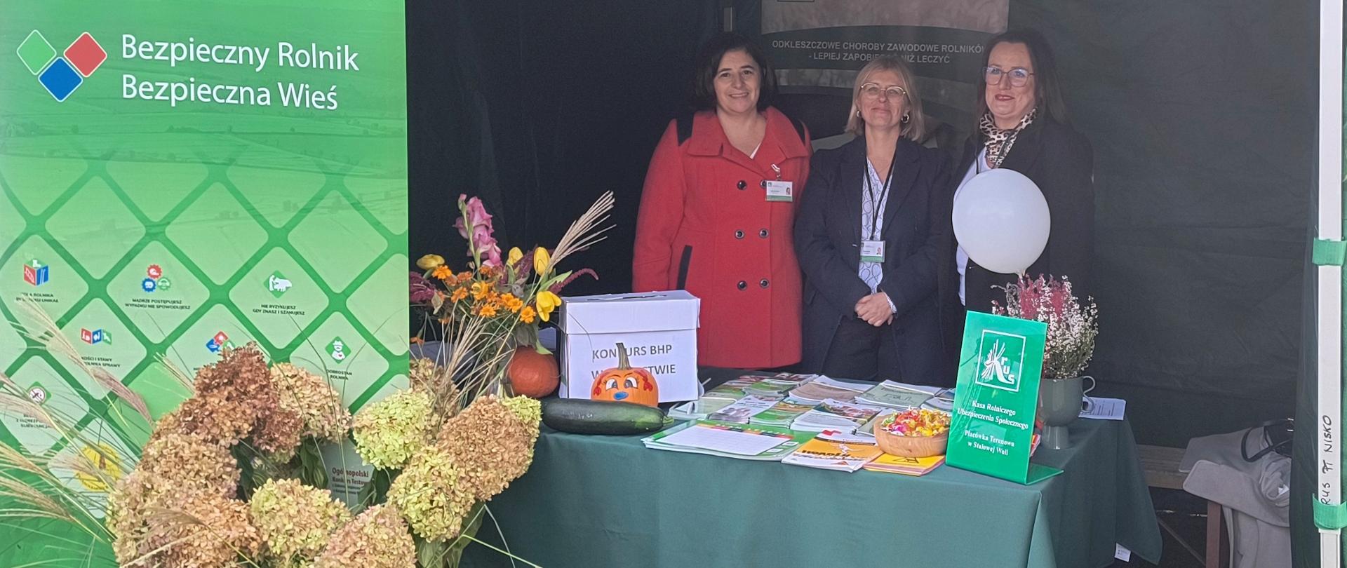 Przed trzema kobietami stojącymi przy stoisku znajduje się stół, na którym wyłożone są materiały prewencyjne Kasy (ulotki, opaski odblaskowe) oraz roll-up z hasłem:” Bezpieczny Rolnik Bezpieczna Wieś”. 