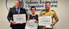 Dwaj mężczyźni i jedna kobieta prezentują bony z kwotami dotacji stojąc na tle białej ściany i napisem Wojewódzki Fundusz Ochrony Środowiska i Gospodarki Wodnej w Warszawie. 