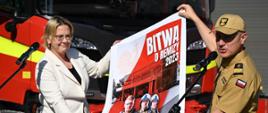 Kobieta oraz strażak w mundurze w kolorze musztardowym trzymają plakat na którym jest napis bitwa o remizy o prezentują go. 