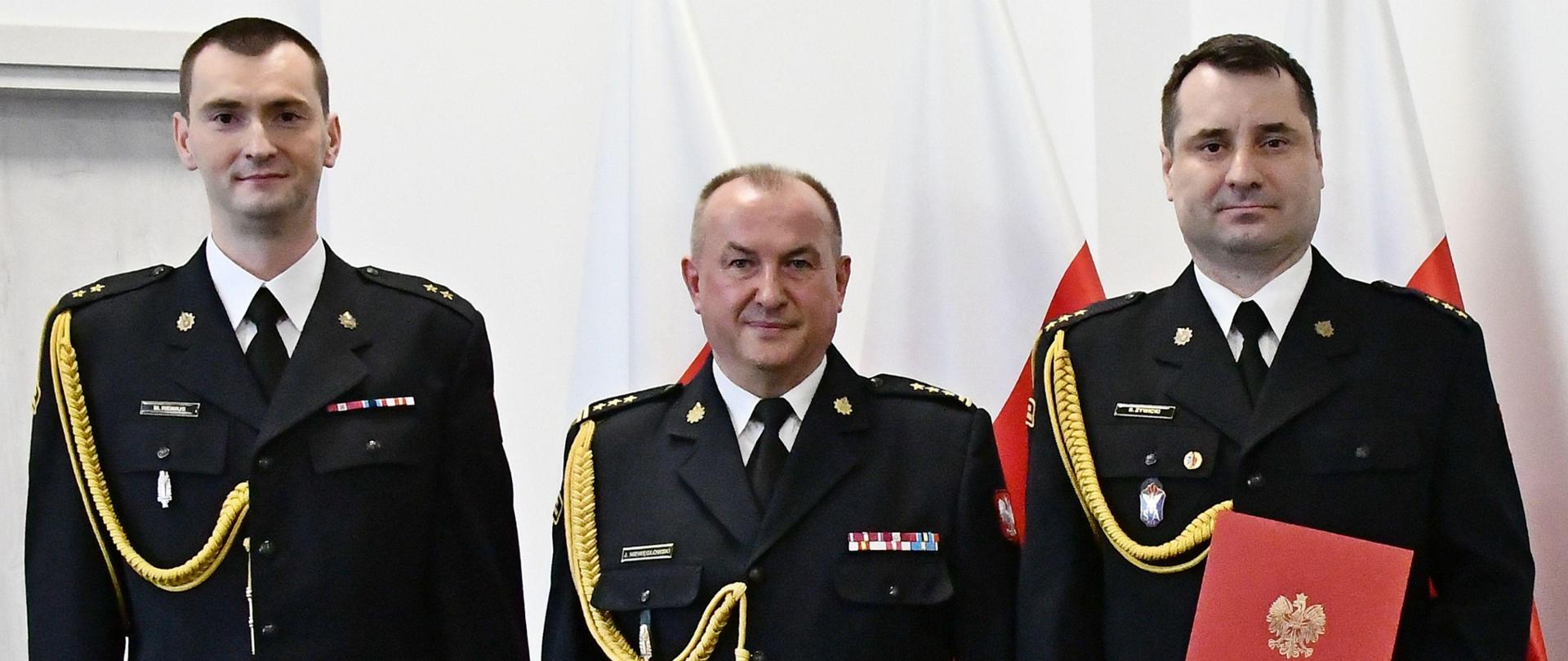 Trzech strażaków Państwowej Straży Pożarnej w mundurach wyjściowych ze sznurem stoi obok siebie jeden strażak trzyma czerwoną teczkę za nimi ustawione są trzy flagi Polski oraz znajdują się drzwi. 