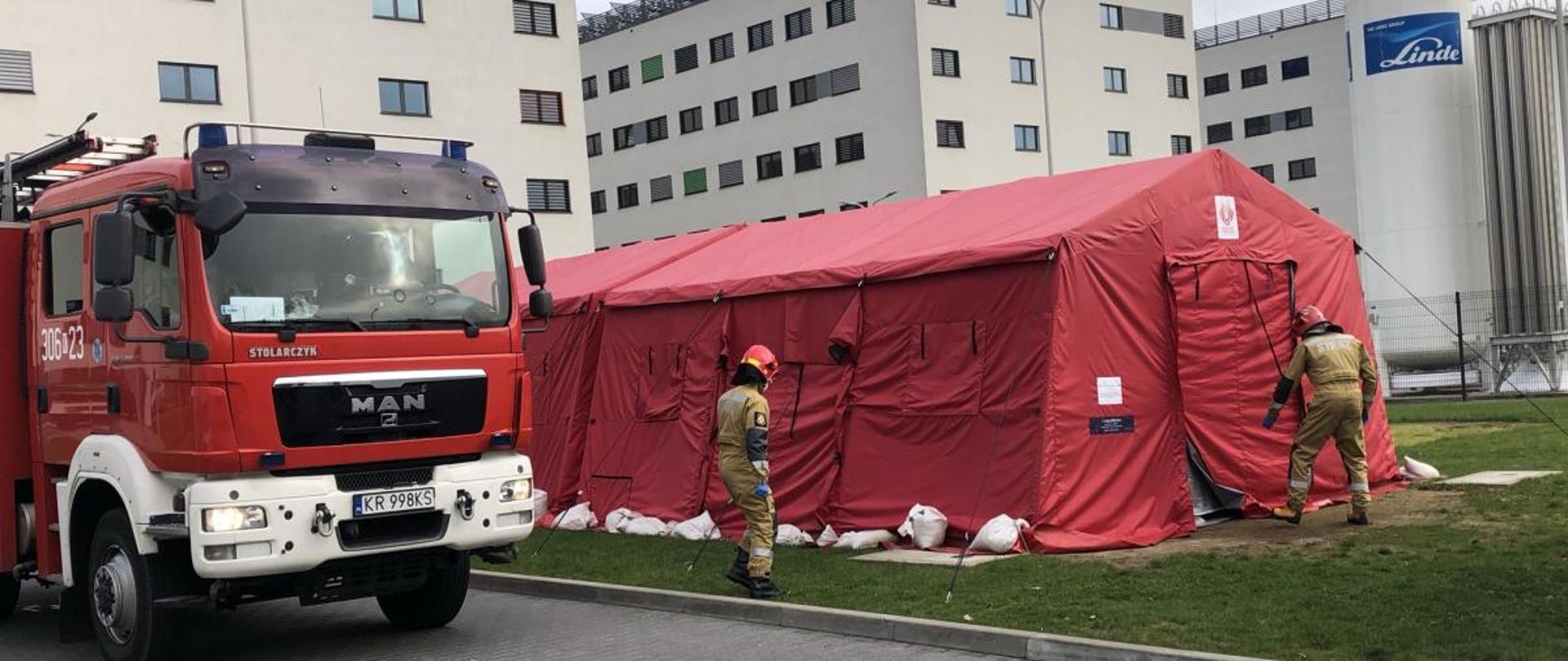 Po lewej stronie wóz strażacki, po prawej rozłożony czerwony duży namiot, w oddali budynki wielopiętrowe. Przed namiotem dwóch strażaków. 