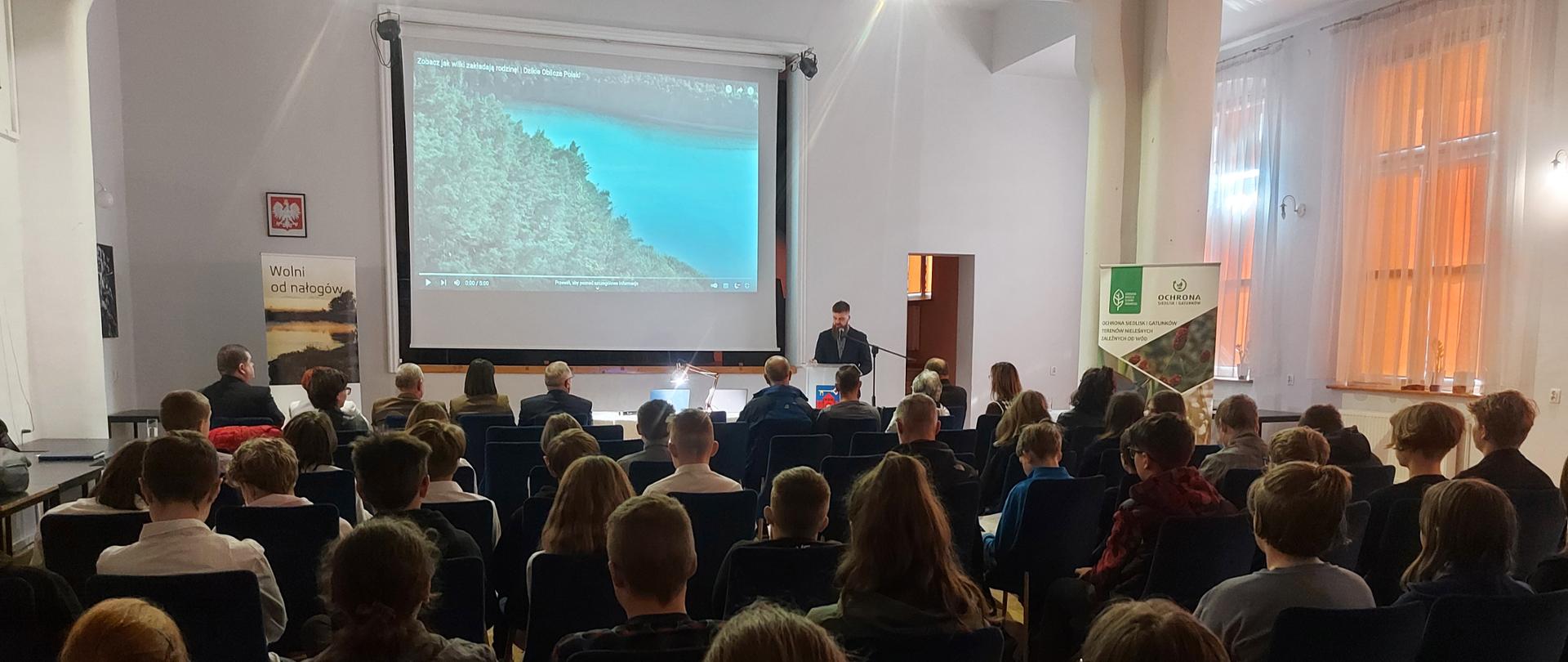 Wnętrze sali, w której na krzesłach odwrócona tyłem siedzi grupa kilkudziesięciu osób. W oddali na ścianie wisi ekran z wyświetlonym filmem, po prawem stronie stoi mężczyzna, przemawiający do mikrofonu.