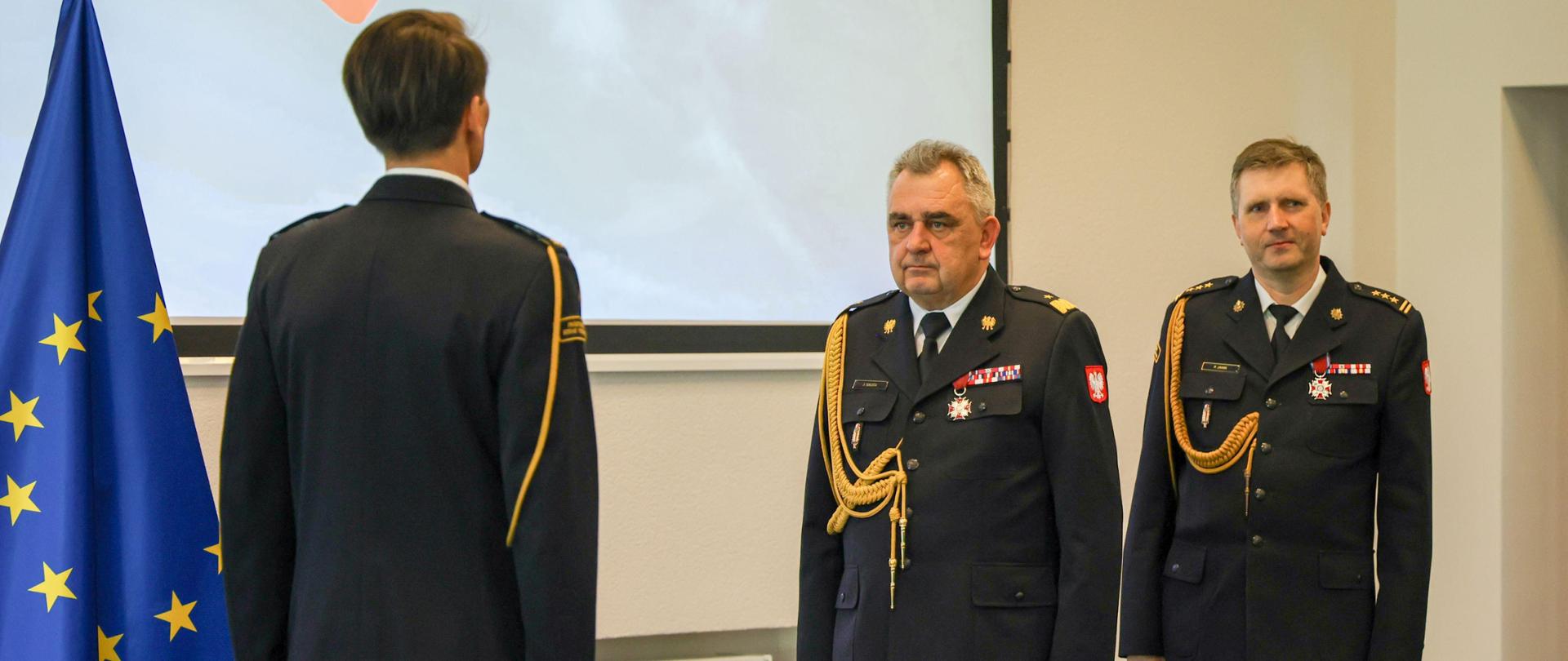 Zastępca komendanta głównego PSP przyjmuje meldunek od dowódcy uroczystości obchodów dnia strażaka w Centrum Naukowo-Badawczym Ochrony Przeciwpożarowej im. Józefa Tuliszkowskiego Państwowym Instytucie Badawczym w Józefowie. Widoczna flaga Unii Europejskiej.