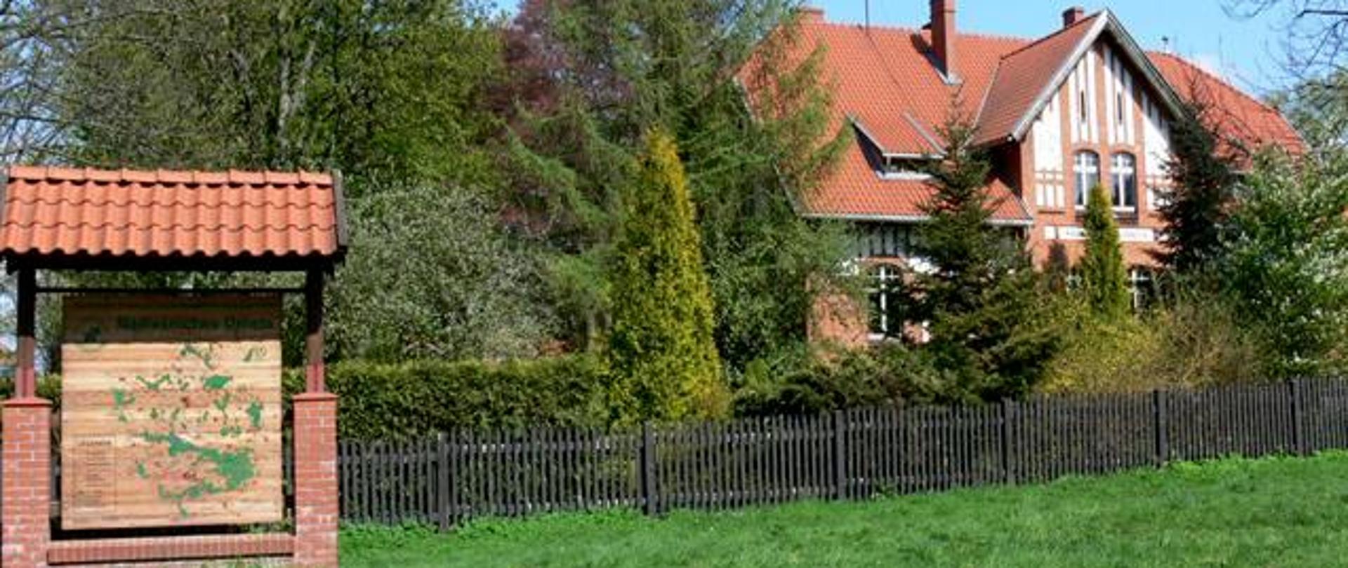 Budynek - siedziba nadleśnictwa z czerwonej cegły, kryty dachówką, w lewym dolnym rogu tablica informacyjna z mapa, przed budynkiem rosną drzewa: świerkowe, modrzew i krzewy ozdobne.