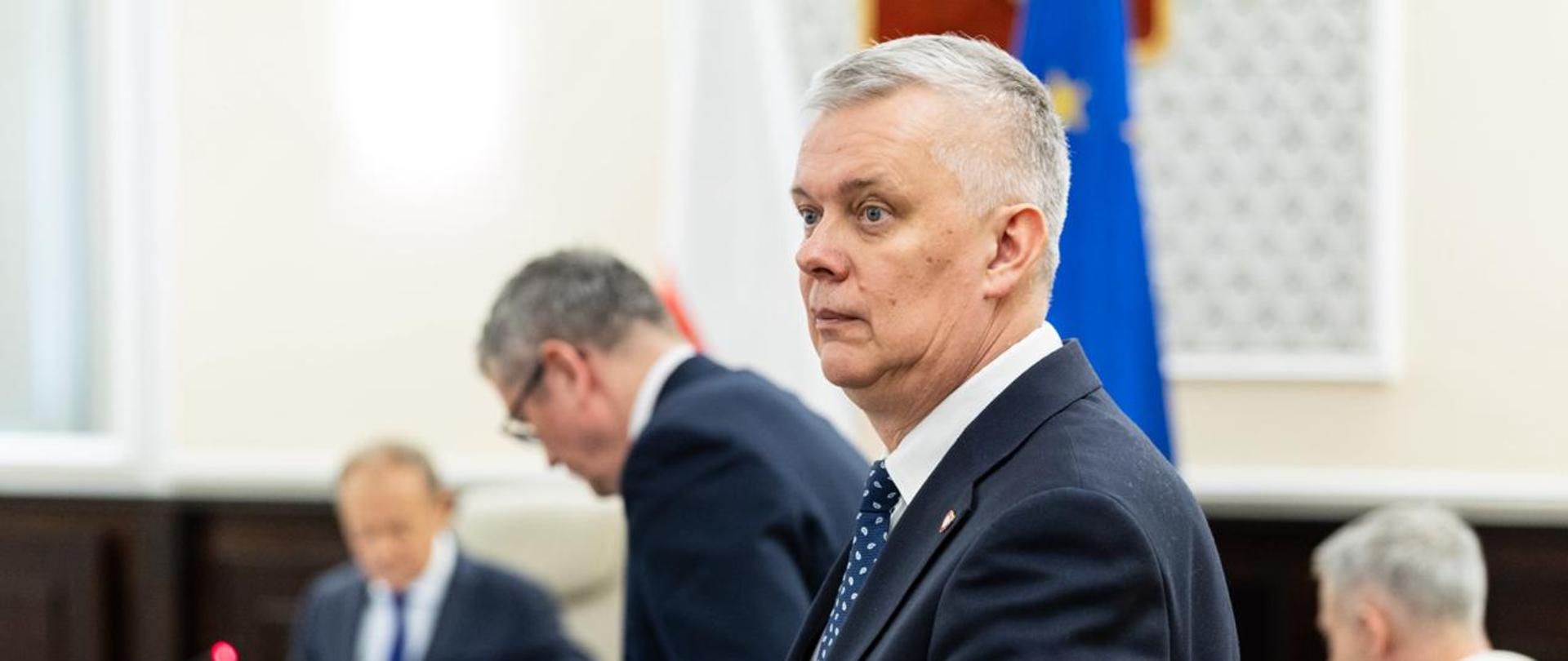 Na pierwszym planie widać stojącego ministra Tomasza Siemoniaka. Minister trzyma ręce na fotelu. W tle widać trzy inne osoby m.in. siedzącego tyłem po prawej stronie ministra Gawkowskiego oraz po lewej stronie zdjęcia siedzącego premiera Donalda Tuska. Po lewej stronie widać również stół, na którym znajdują się monitory, termosy, butelki z wodą i sokami, mikrofony. Za premierem Donaldem Tuskiem znajdują się dwie flagi - po prawej flaga UE (niebieska z żółtymi gwiazdkami) oraz polska (biało-czerwona). 