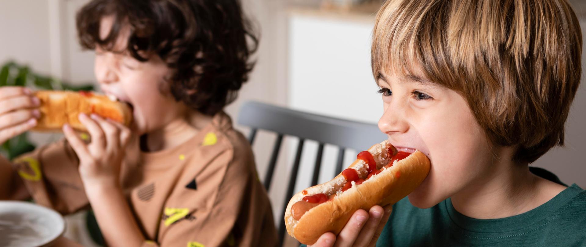 Pomieszczenie, dwoje dzieci przy stole je hot-dogi.