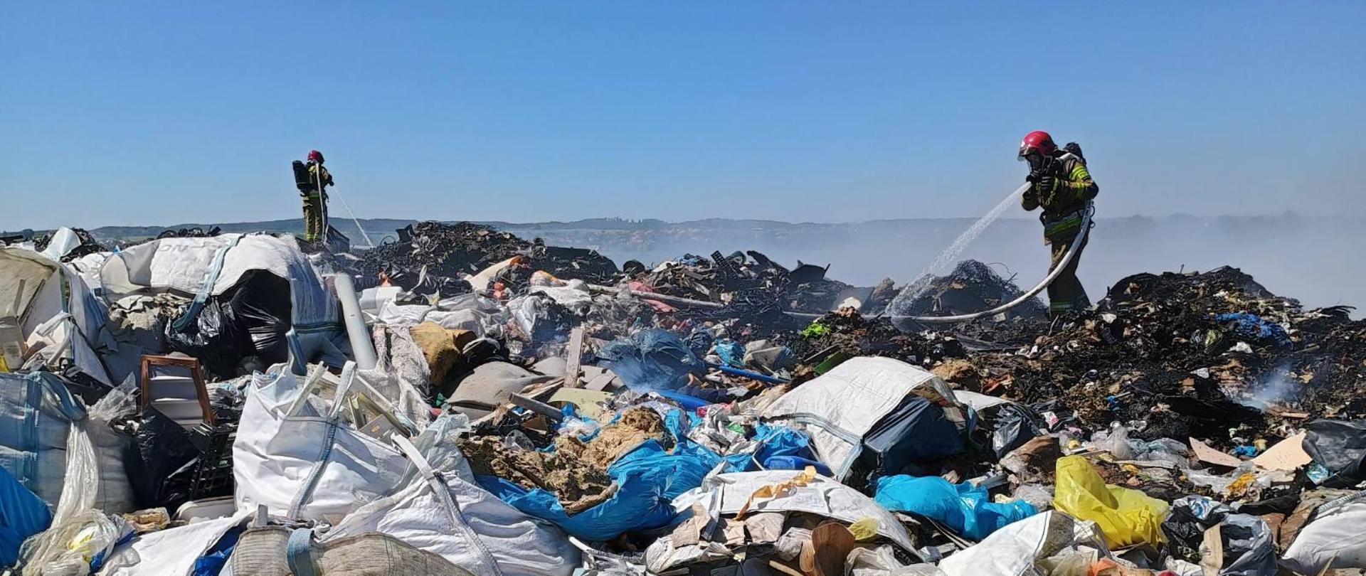 sterty płonących śmieci, na górze strażacy gaszący ogień
