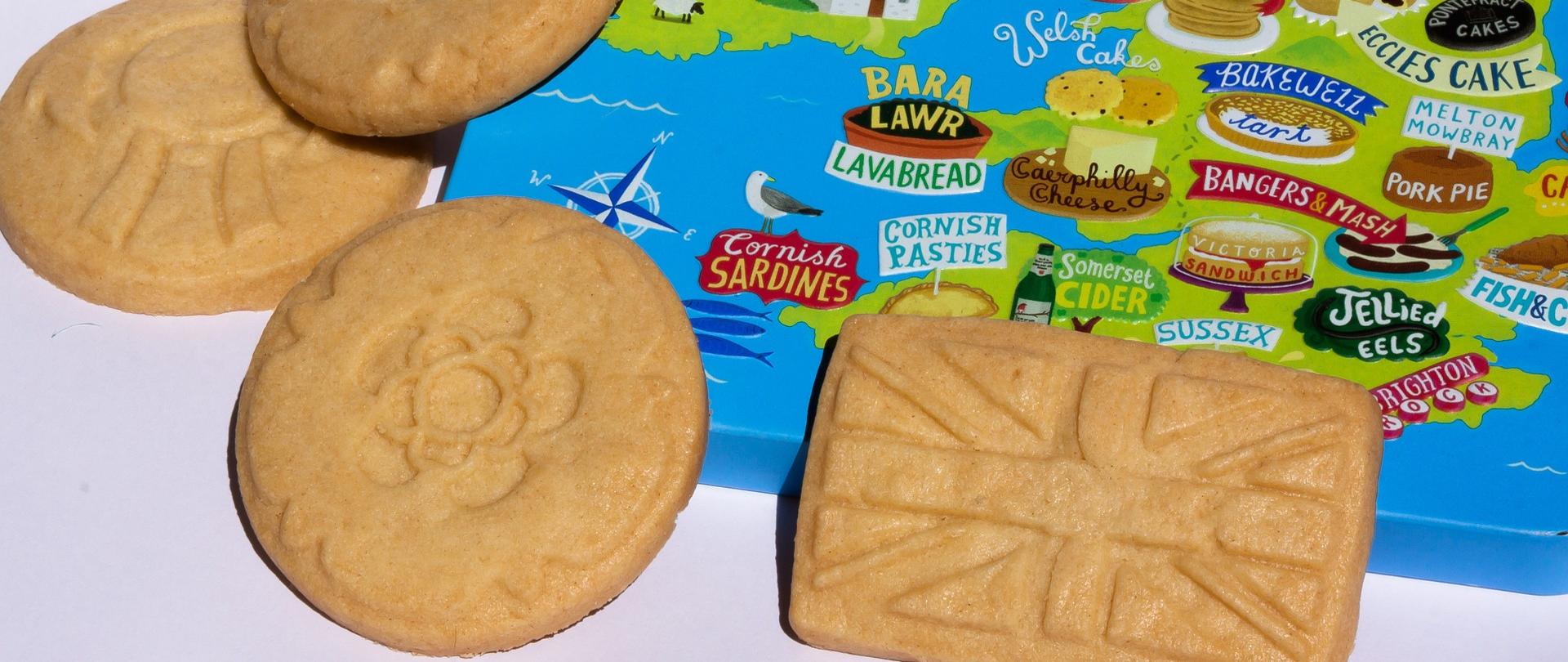 Na pierwszym planie znajdują się cztery ciasteczka: pierwsze z prawej prostokątne ma wzór flagi brytyjskiej, trzy pozostałe są okrągłe z kształtami kwiatów. Na drugim planie znajduje się graficzna mapa Wielkiej Brytanii.