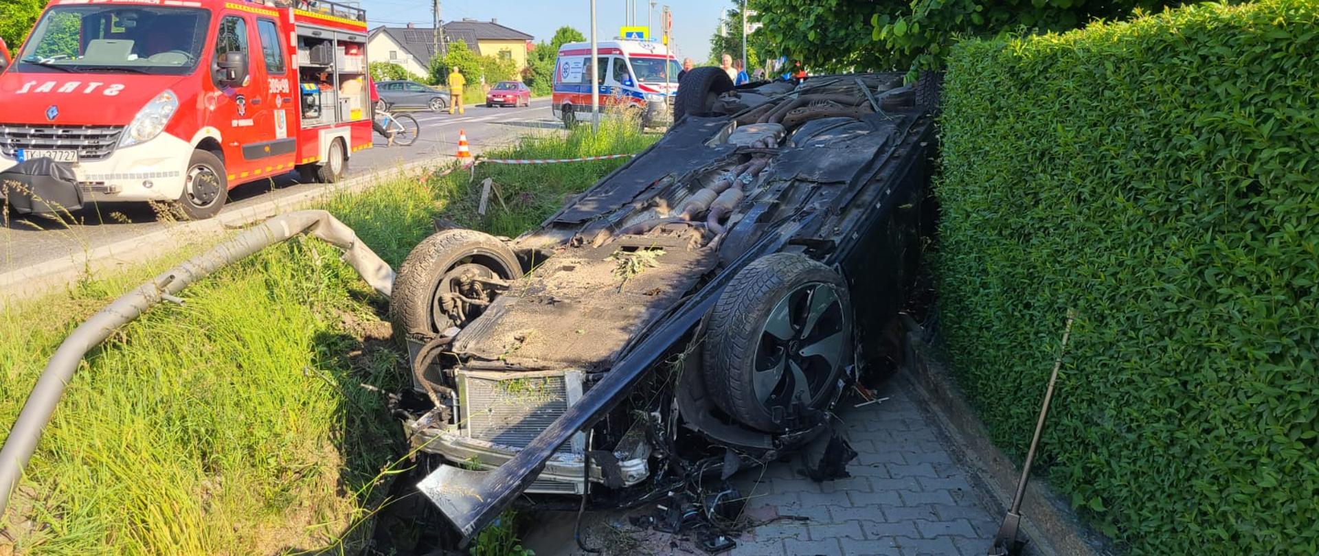Zdjęcie przedstawia samochód Audi leżący na dachu na chodniku. Z lewej strony leży zniszczona latarnia uliczna , widać także ambulans i wóz strażacki